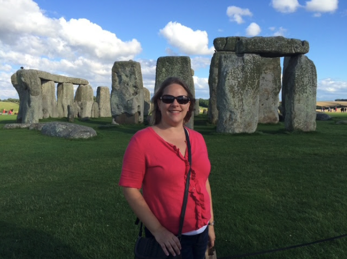A woman in a pink top wearing sunglasses, smiling in front of Stonehenge on a sunny day with blue skies and green grass.